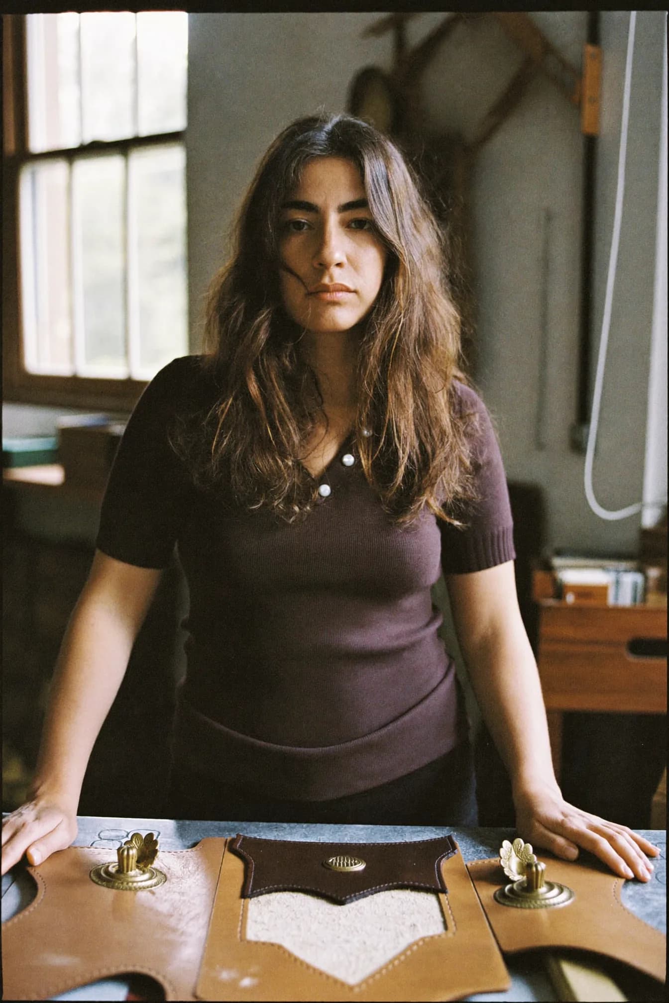 Inês Carvalho at a leather-cutting bench, laying a steel clicker die onto a hide, with finished belts hanging on a rail behind her.