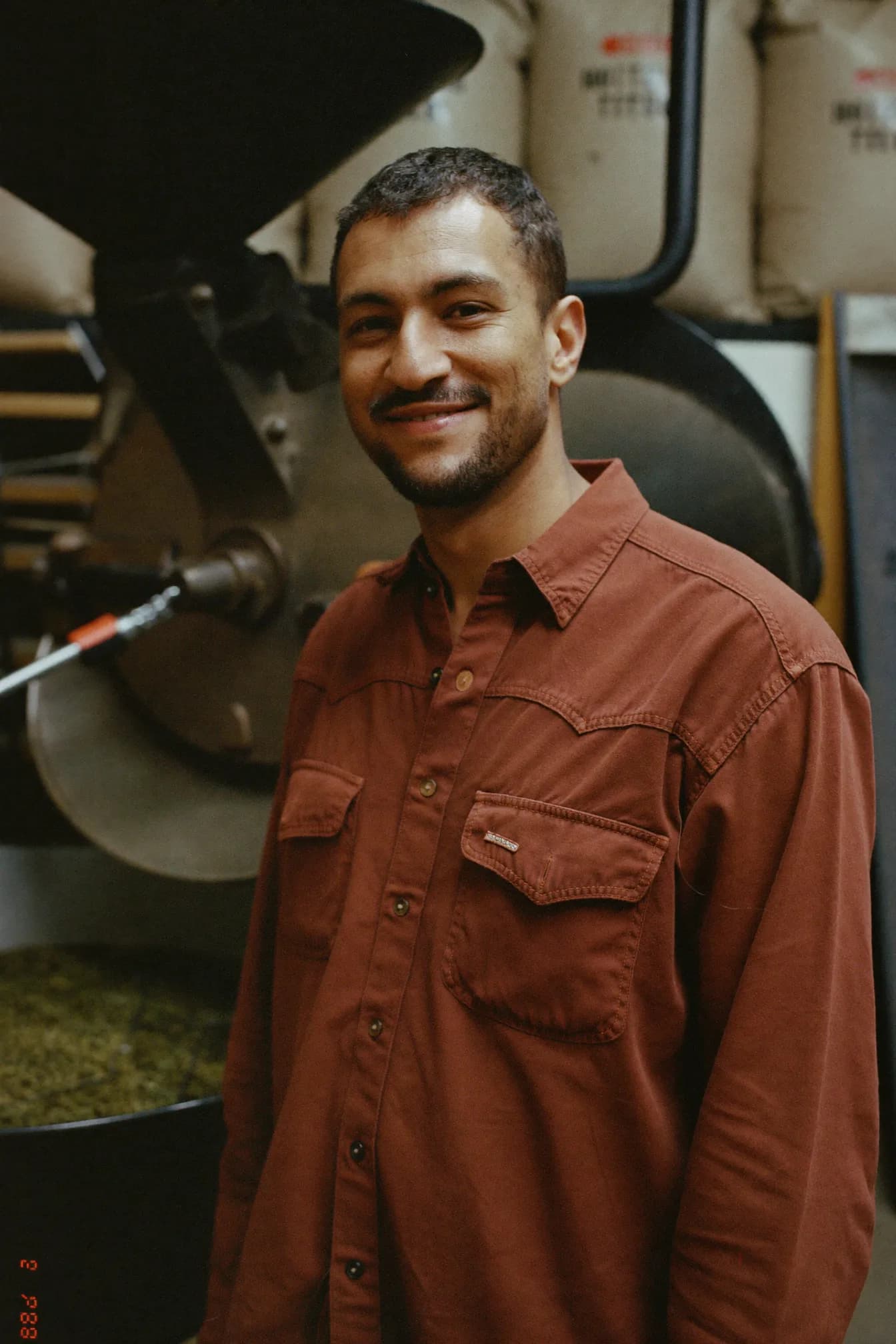 Marcus Reeve beside a drum roaster, pulling a sample tray of beans, with stacked sacks of green coffee behind him.