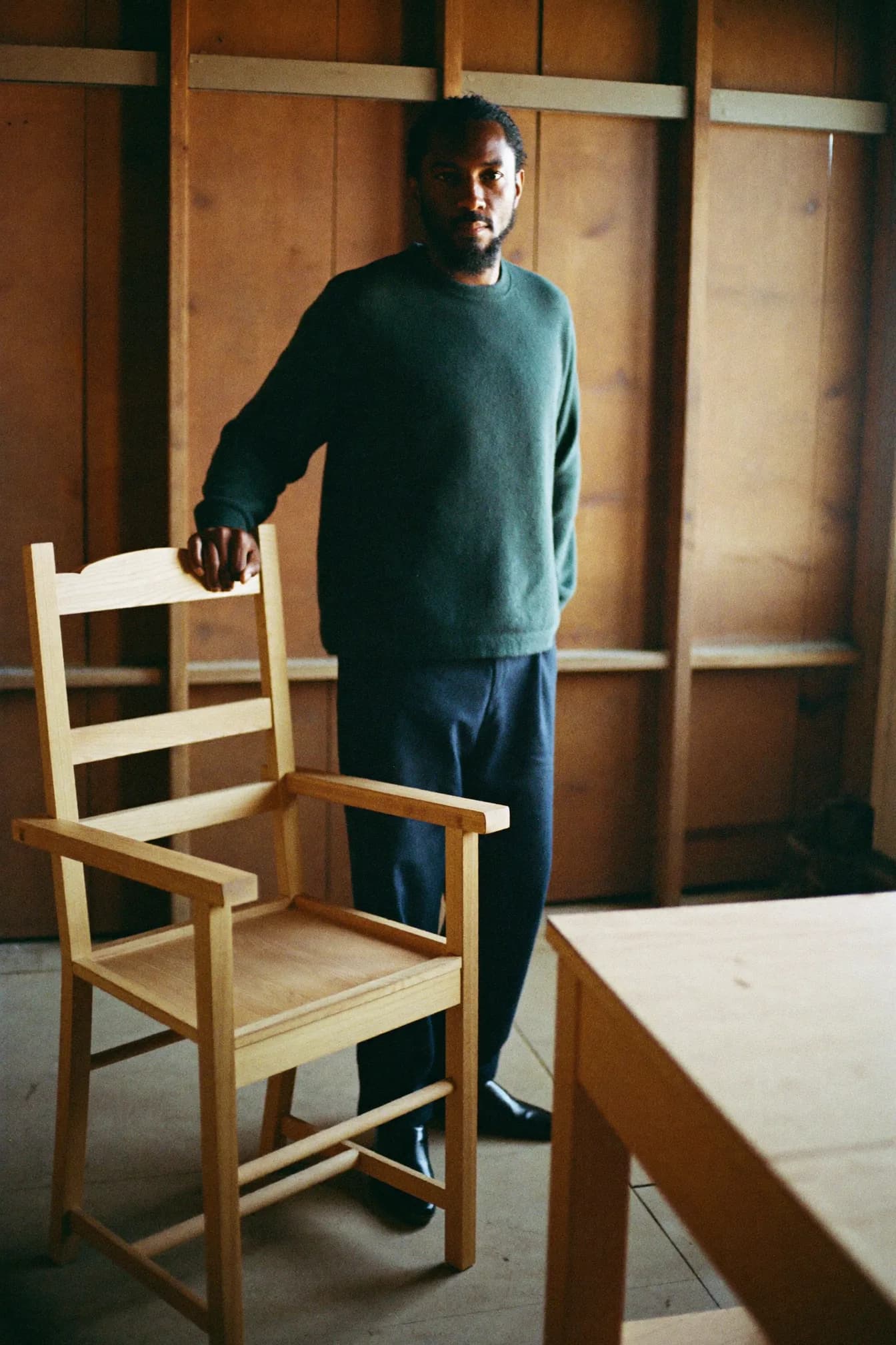 Martin Oduya in the timber yard at Brackwood, one hand resting on a stack of seasoning oak boards separated by thin battens.
