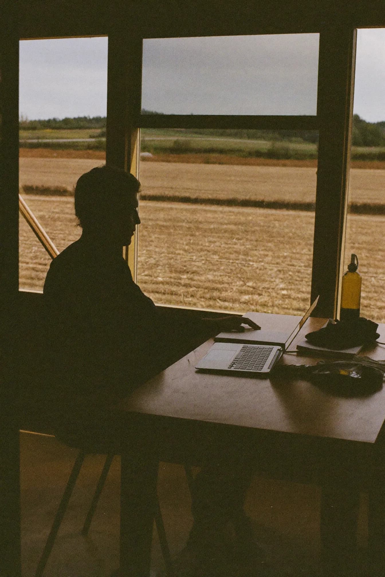 Sam Devlin at a desk in a wood-panelled co-op office, a laptop open beside a spiral notebook and a two-way radio.