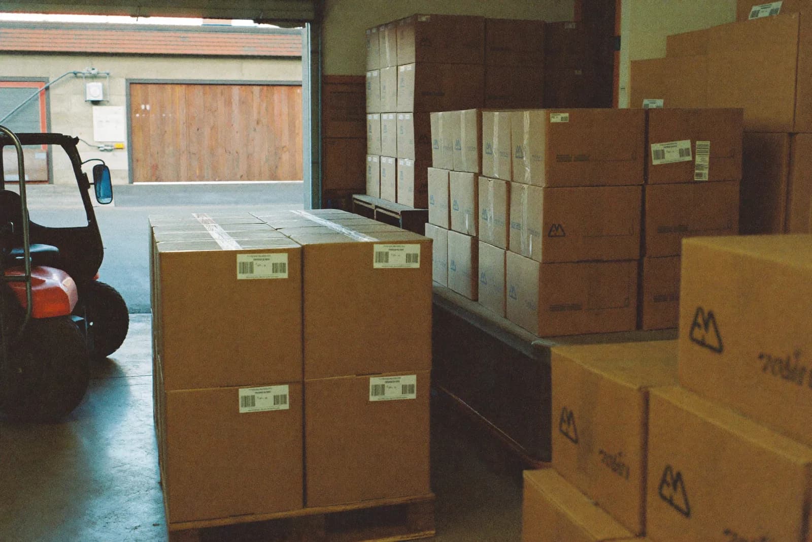 A small warehouse with pallets of components stacked to head height and a roller door open to a loading yard.