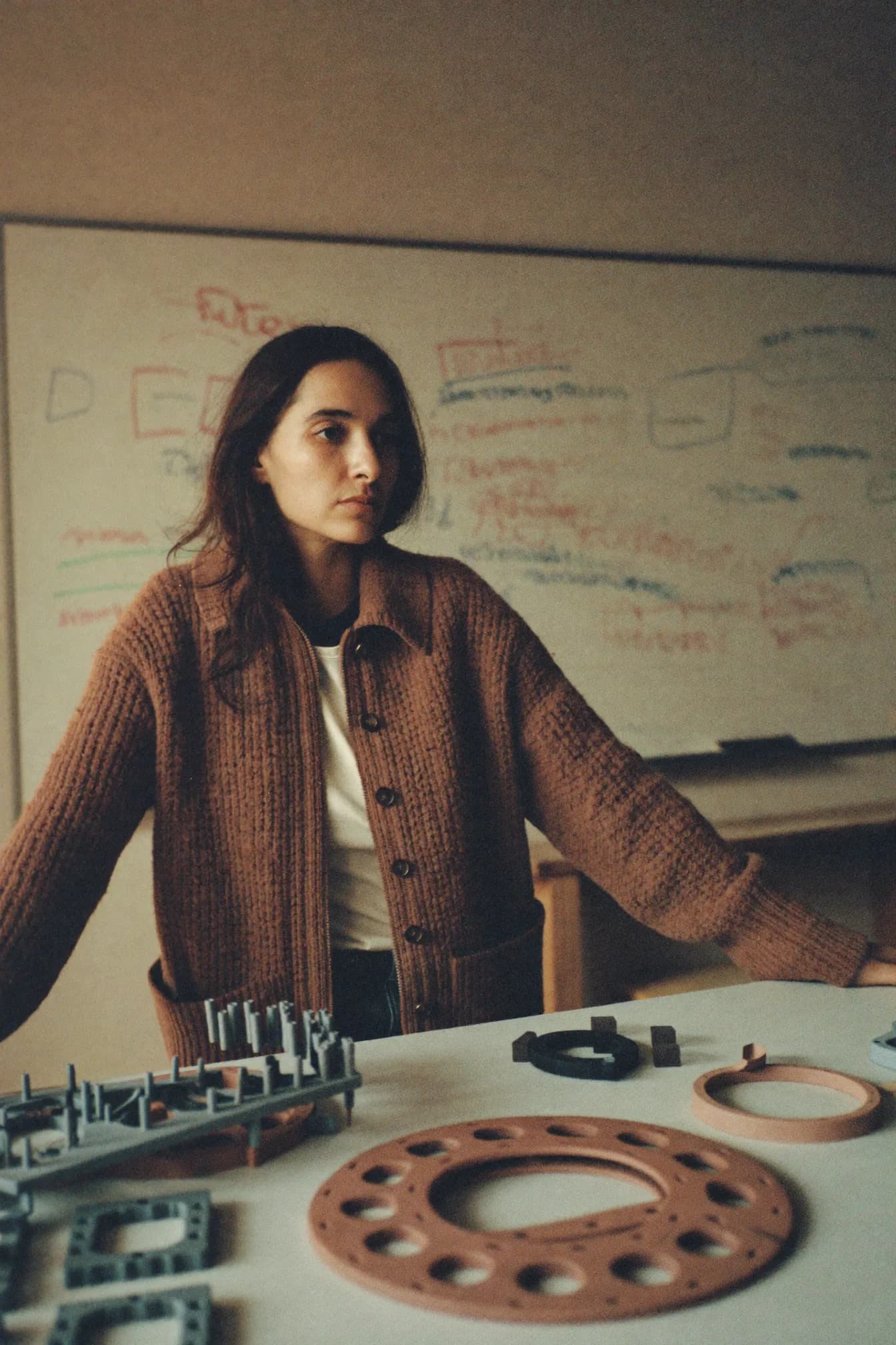 Ana Ferreira standing at a work table covered with moulded prototype housings, a whiteboard of red and green diagrams behind her.
