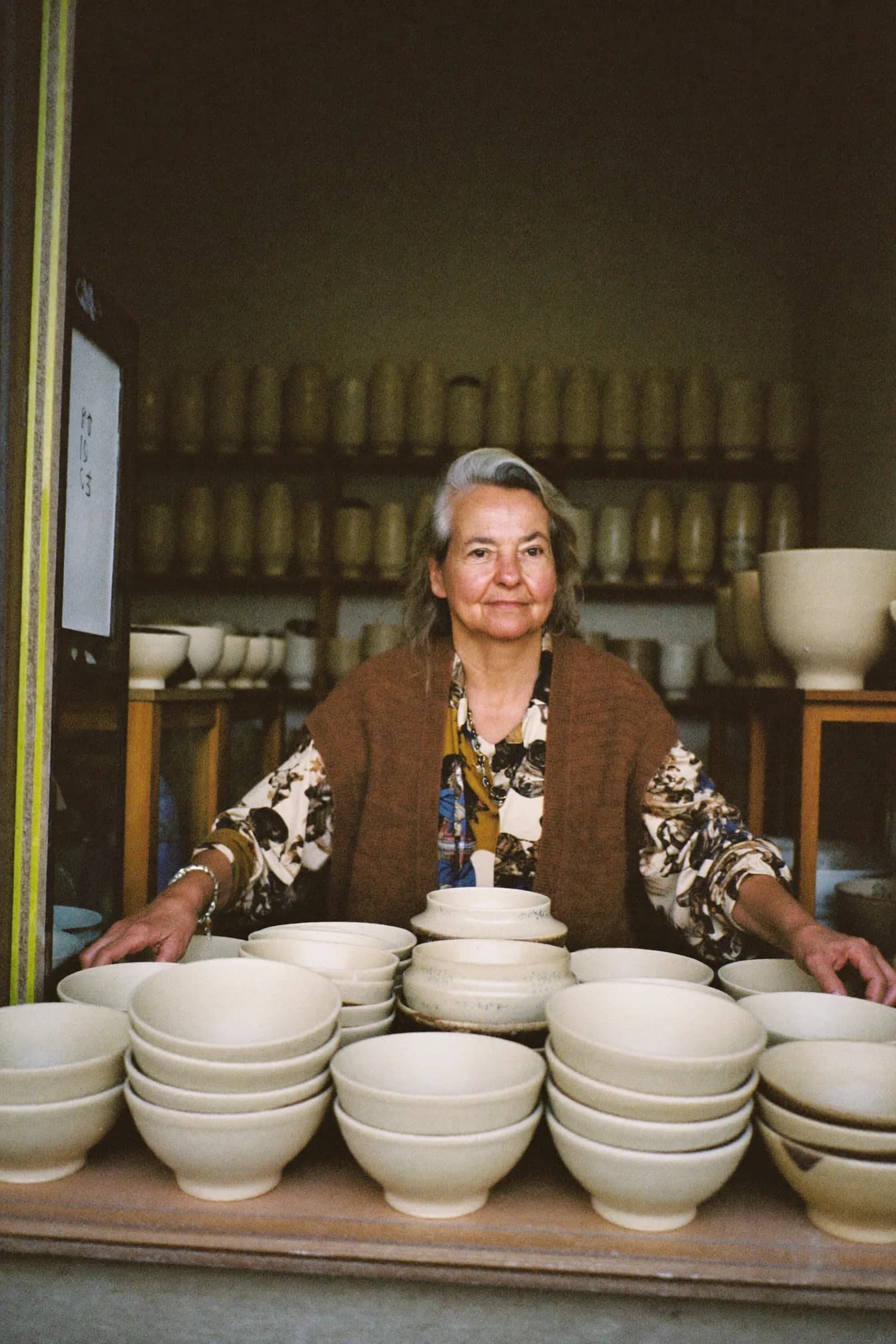 Della Marsh at a potter’s wheel positioned in her shop window, centring a bowl, with shelves of glazed pieces visible behind her.