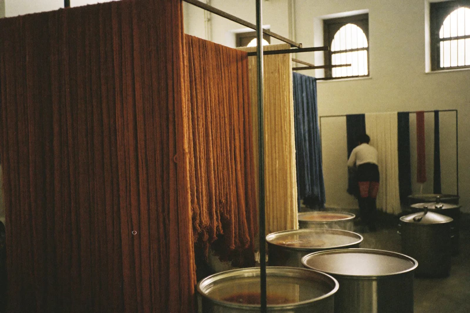 A dye house interior with steel vats, hanging skeins of rust and indigo yarn, and a wet concrete floor.