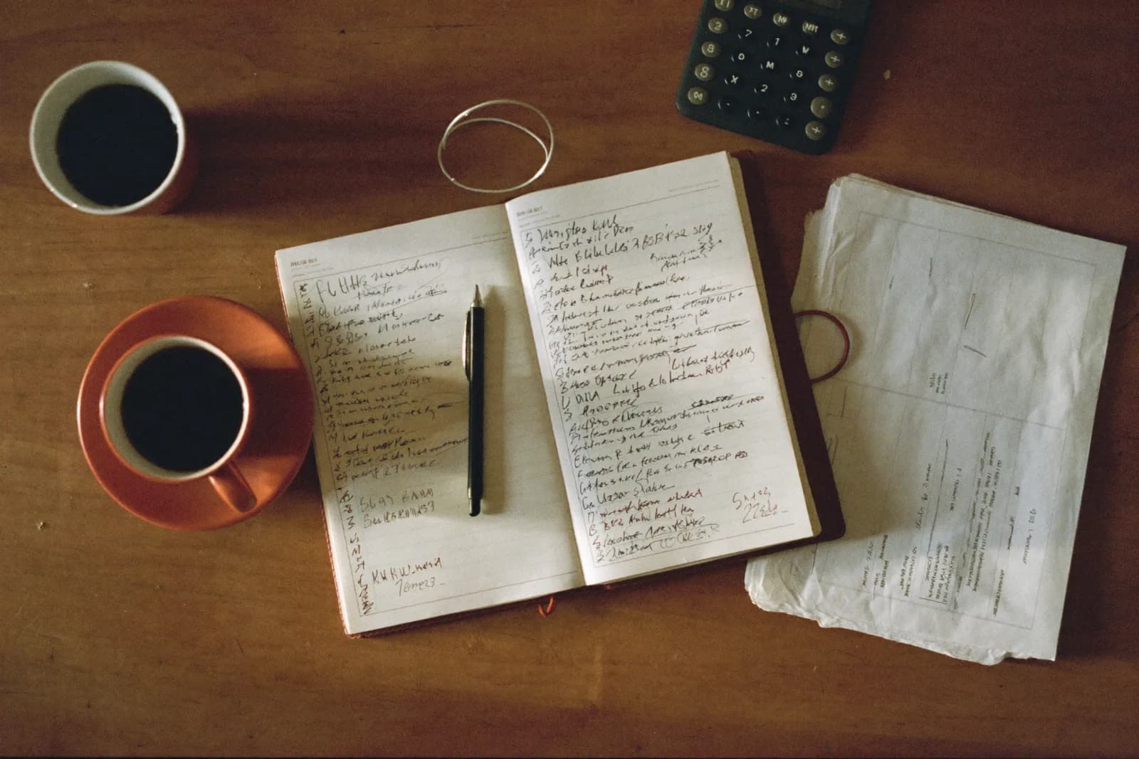 A ledger book open on a desk beside a pen, calculator and stacked invoices.