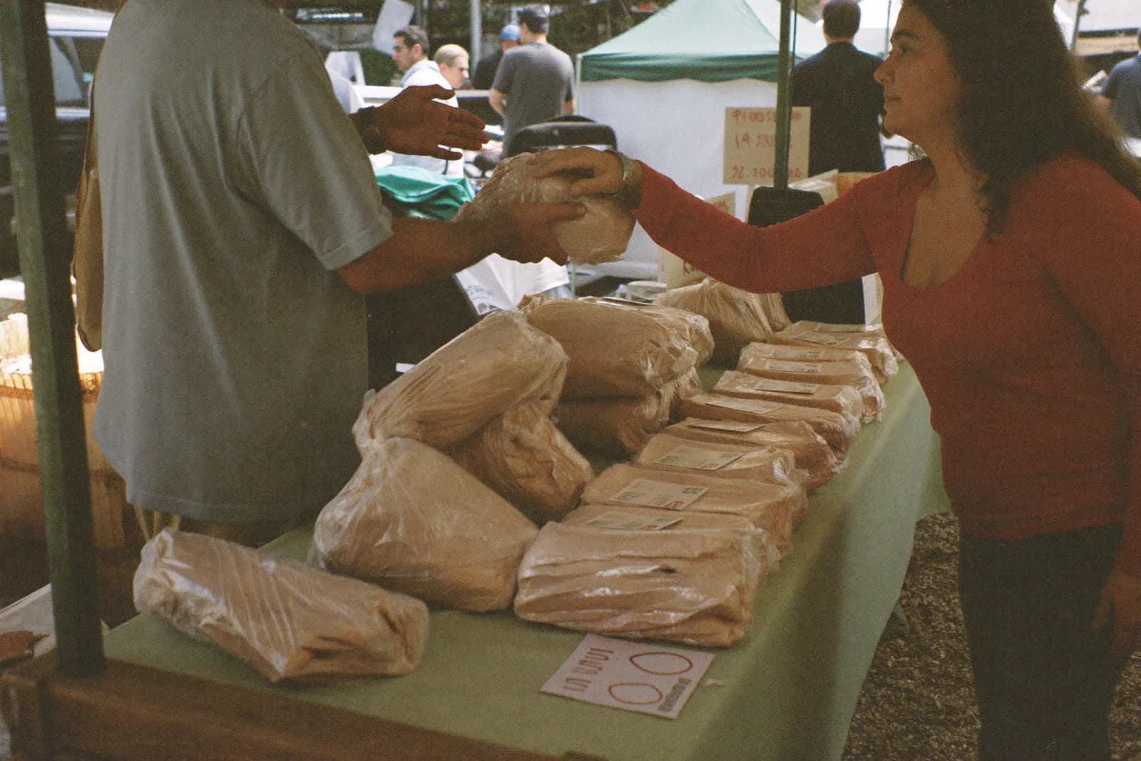 A trestle table at an outdoor market, stacked with goods, a vendor talking to a passer-by.