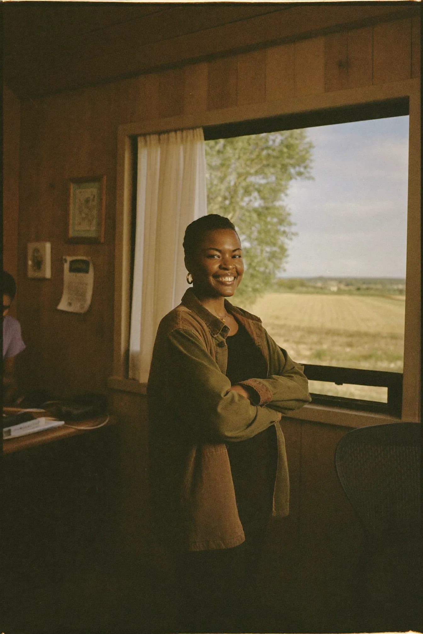 Maya Okonjo leaning against a windowsill in a wood-panelled co-op office, smiling, with a harvested field visible through the window behind her.